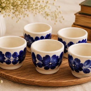 Peut inclure: Un ensemble de cinq petites tasses en céramique blanc cassé disposées sur un plateau rond en bois. Chaque tasse présente un motif peint à la main de fleurs et de feuilles bleu foncé entourant l'extérieur. Les tasses ont un bord lisse.