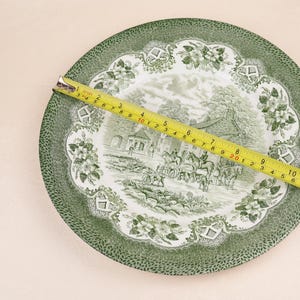 May include: A green and white decorative plate with a landscape scene and floral border. A yellow tape measure is laid across the plate, showing a measurement of approximately 11 inches.