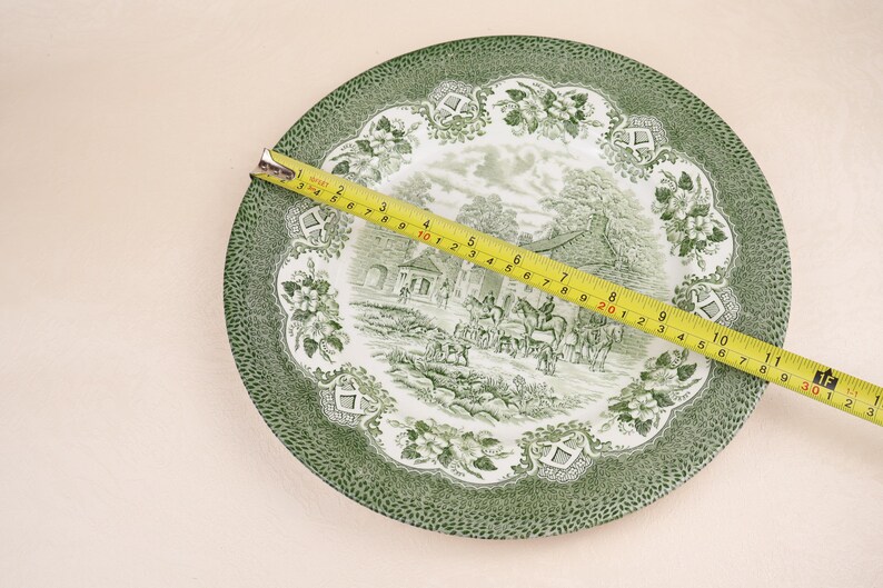 May include: A green and white decorative plate with a landscape scene and floral border. A yellow tape measure is laid across the plate, showing a measurement of approximately 11 inches.