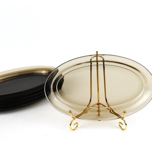 May include: A stack of oval-shaped, dark-tinted glass serving platters. One platter is displayed on a gold-colored metal stand. The platters are a smoky, translucent color, and the stand has a decorative design.
