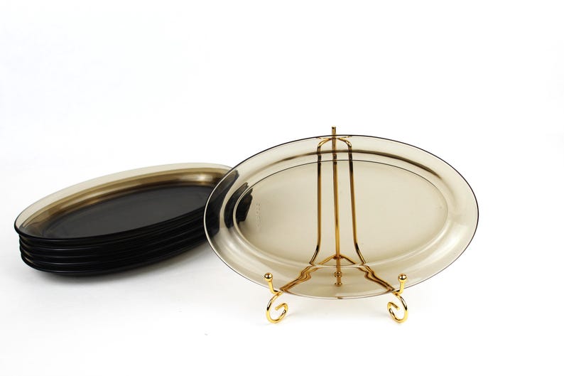 May include: A stack of oval-shaped, dark-tinted glass serving platters. One platter is displayed on a gold-colored metal stand. The platters are a smoky, translucent color, and the stand has a decorative design.