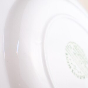 May include: Close-up of a white ceramic plate with a curved rim. The plate has a glossy finish and a subtle, curved design. A green and white stamp is visible on the bottom right corner.