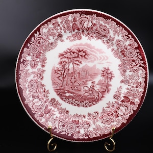 May include: A round, white and burgundy plate with a detailed floral border and a central landscape scene. The plate is displayed on a gold-colored stand, showcasing its intricate design and vintage aesthetic.