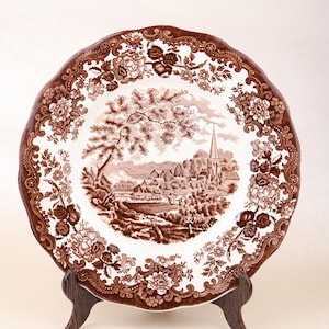 May include: A brown and white ceramic plate with a detailed landscape scene and floral border. The plate is displayed on a wooden stand. The landscape features a church, trees, and a village.