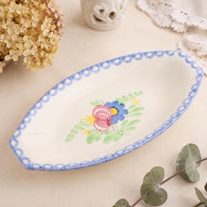 May include: Oval-shaped, off-white ceramic serving dish with a blue scalloped border. The dish features a hand-painted floral design in the center with pink, blue, and yellow flowers and green leaves. A decorative piece for serving food.