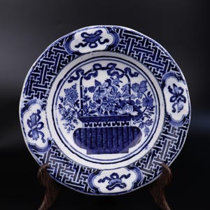 May include: A blue and white porcelain plate featuring a floral basket design. The plate has a patterned border and is displayed on a wooden stand. The design is a classic example of blue and white pottery, a traditional style.