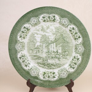 May include: A green and white decorative plate with a detailed landscape scene featuring buildings, trees, and figures on horseback. The plate has a textured green border and floral accents, displayed on a wooden stand.