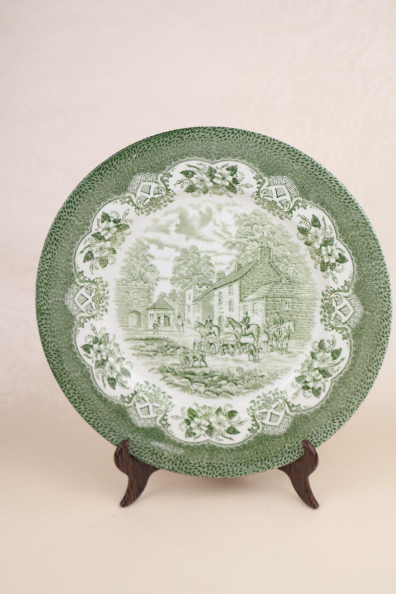 May include: A green and white decorative plate with a detailed landscape scene featuring buildings, trees, and figures on horseback. The plate has a textured green border and floral accents, displayed on a wooden stand.