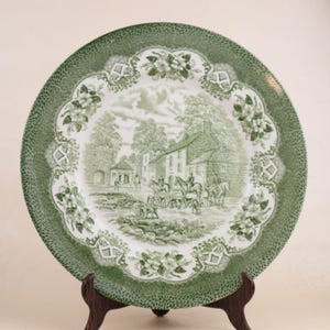 May include: A green and white decorative plate with a detailed landscape scene. The plate features a green border with a dotted pattern and a central design of a village scene with figures and buildings. The plate is displayed on a wooden stand.