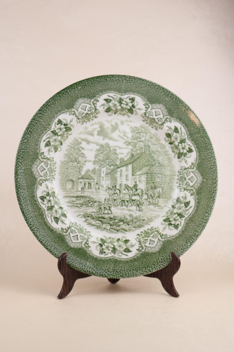 May include: A green and white decorative plate with a detailed landscape scene. The plate features a green border with a dotted pattern and a central design of a village scene with figures and buildings. The plate is displayed on a wooden stand.