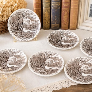 Op de afbeelding: Een set van zes witte keramische borden met een bruin pastorale tafereel ontwerp. De borden zijn gerangschikt op een wit kanten tafelkleed, met een groter bord op een gouden standaard. Decoratieve boeken en een ingelijste foto op de achtergrond.