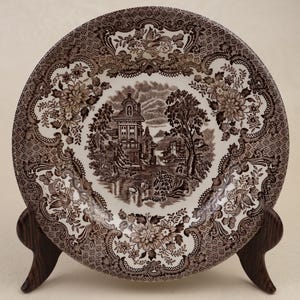 Könnte beinhalten: Ein runder Keramikteller mit einem detaillierten braun-weißen Transferdruckmuster. Die Mitte zeigt eine Landschaft mit einem Gebäude und Bäumen, während der Rand mit komplexen Blumenmotiven und geometrischen Mustern verziert ist.