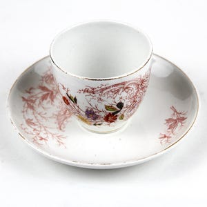 Könnte beinhalten: Ein weißes Tee-Set, bestehend aus Tasse und Untertasse. Die Tasse und Untertasse haben ein Blumen- und Blätterdesign in Rot-, Braun- und Grüntönen. Die Ränder der Tasse und Untertasse sind goldfarben. Das Set steht auf weißem Hintergrund.