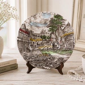 May include: A round decorative ceramic plate featuring a detailed transferware scene of a rural English village with thatched-roof cottages, a stone castle in the distance, figures, and a small dog, rendered in shades of black, green, and yellow.