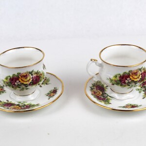 May include: Two white teacups and saucers with a floral rose pattern in yellow, red, and green. The rims and edges are trimmed with gold. The teacups have a small handle.
