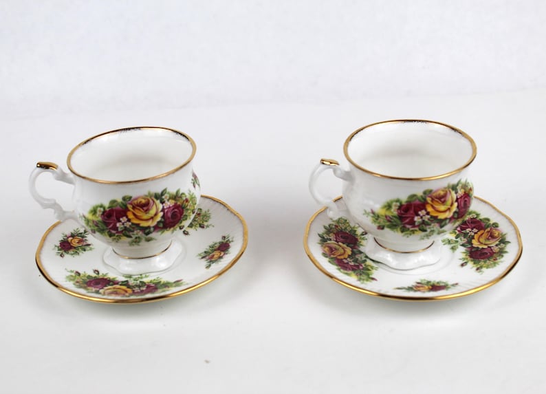 May include: Two white teacups and saucers with a floral rose pattern in yellow, red, and green. The rims and edges are trimmed with gold. The teacups have a small handle.
