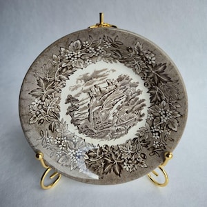 May include: A decorative plate with a brown and white landscape scene, featuring a castle on a cliff, surrounded by trees. The plate has a floral border and is displayed on a gold-coloured stand. The plate is approximately 20 cm in diameter.