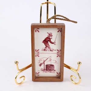May include: A small, rectangular wooden frame with a decorative tile featuring a red illustration of a person fishing. The frame is suspended by a rope and displayed on a gold-coloured easel.