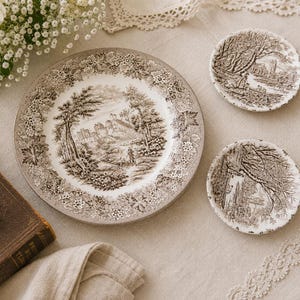 May include: A set of vintage-style brown and white ceramic plates and saucers. The large plate features a landscape scene with a castle, trees, and a figure. Two smaller saucers have similar landscape designs. The plates are surrounded by a floral border.