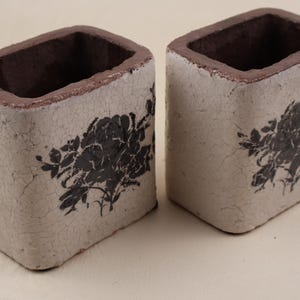Peut inclure: Deux récipients rectangulaires avec une finition extérieure blanc cassé craquelée et un bord intérieur marron foncé. Chaque vase présente une impression graphique florale noire sur la face avant, illustrant un motif botanique détaillé.