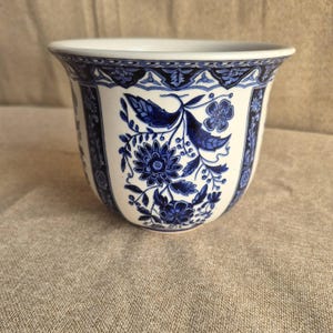 May include: A white ceramic planter with a blue and white floral design. The planter has a wide mouth and a rounded bottom.