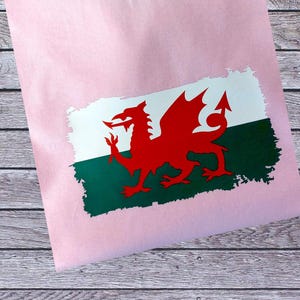以下が含まれることがあります： ウェールズの国旗のデザインが施された、明るいピンク色のTシャツ。旗は、緑のストライプの上に白い背景に赤いドラゴンが描かれています。Tシャツは木製の表面に折りたたまれています。