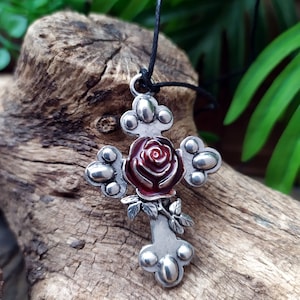 Könnte beinhalten: Ein silberner Kreuzanhänger mit einer roten Rose in der Mitte. Das Kreuz hat ein detailliertes Design mit kleinen Kreisen an den Rändern. Der Anhänger hängt an einer schwarzen Schnur.