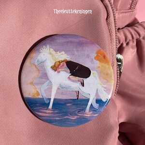 Op de afbeelding: Een ronde button met een illustratie van een persoon met roodachtig haar in een donkere top, liggend op de rug van een wit paard in blauw water. De tekst "Theeleuttekeningen" staat in het wit boven het paard afgebeeld.