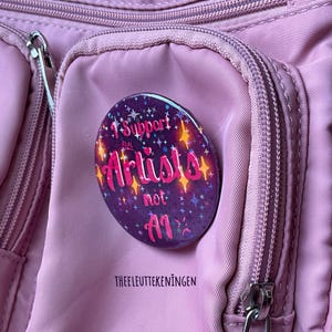 Può includere: Un bottone viola appuntato a una borsa rosa. Il bottone riporta la scritta "I Support Real Artists not AI" in rosa e bianco, con stelle e brillantini. La borsa ha più tasche e cerniere.