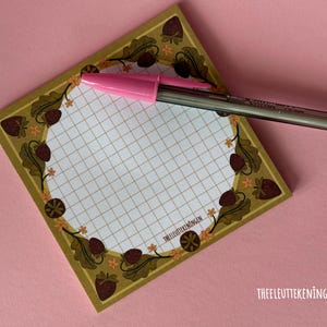 May include: A square notepad with a grid pattern and a decorative border featuring acorns, strawberries, and leaves. A pink pen rests on the notepad. The notepad is on a pink surface. The text "THEELEUTTEKENINGEN" is printed on the notepad.