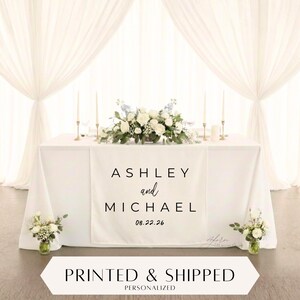 Peut inclure: Une table blanche ornée d'une composition florale et d'un chemin de table personnalisé. Le chemin de table affiche les noms "Ashley et Michael" et la date "08.22.26". La table est décorée de bougies et de petits arrangements floraux.