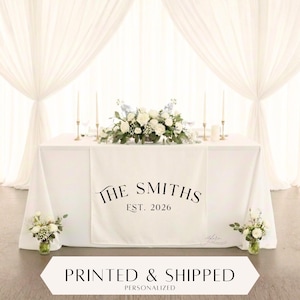 Puede incluir: Una mesa blanca con un camino de mesa personalizado con "The Smiths EST. 2026" en escritura negra. La mesa está decorada con un arreglo floral, velas y pequeños ramos. El fondo incluye cortinas blancas.