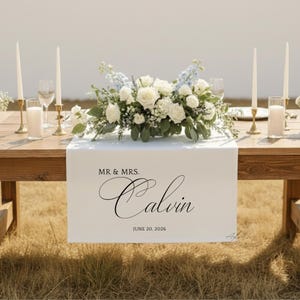 Puede incluir: Una mesa de boda con un camino de mesa blanco con el texto "MR & MRS. Calvin" y la fecha "JUNE 20, 2026". La mesa est&aacute; decorada con un arreglo floral de rosas blancas y flores azules, velas y cristaler&iacute;a.