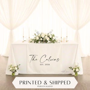 Puede incluir: Una mesa blanca decorada con un centro floral y velas. Un camino de mesa blanco está impreso con "The Calvins EST. 2026". La imagen es de una mesa de recepción de boda. La parte inferior de la imagen tiene el texto "PRINTED & SHIPPED PERSONALIZED".