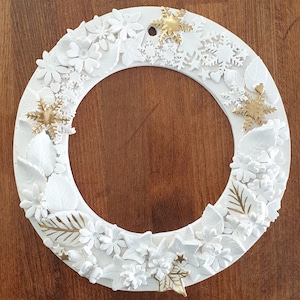 May include: A white wreath decorated with white flowers, leaves, and snowflakes. The wreath is accented with gold glitter stars.