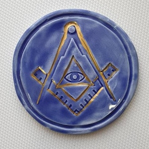 May include: A blue ceramic coaster with a gold-colored all-seeing eye and compass design.