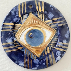 May include: A round ceramic wall hanging featuring a blue and white eye design. The eye is surrounded by a gold diamond shape and gold lines radiating outwards. The background is a dark blue with white speckles.