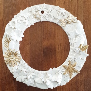 May include: White ceramic wreath with gold accents, featuring snowflakes, flowers, and leaves. The wreath is a decorative piece for a home or office.