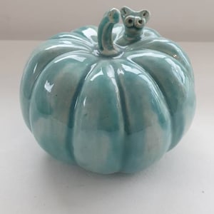 Może przedstawiać: Ceramiczną, turkusową dynia z małą, rzeźbioną twarzą na łodydze.