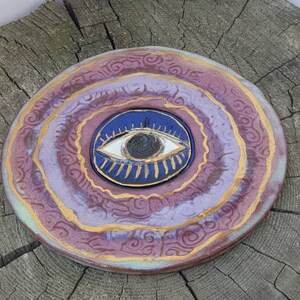 Op de afbeelding: Een keramisch bord met een paars en gouden swirl-ontwerp. In het midden van het bord bevindt zich een blauw oog met een witte pupil en gouden wimpers.