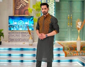 May include: A man wearing a dark gray kurta and matching pants. The kurta features intricate brown embroidery on the chest and collar. He is wearing a watch and black shoes. The background includes a decorative wall and a television.