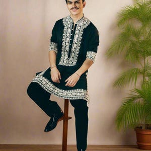 May include: Black tunic and pants set with intricate white embroidery. The tunic features a mandarin collar and button-down front. The outfit is paired with black dress shoes. The person is sitting on a wooden stool next to a potted plant.