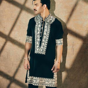 May include: Black tunic with intricate white embroidery. The tunic features a mandarin collar and button-down front. The embroidery details the collar, front, cuffs, and hem. The sleeves are three-quarter length. The person is wearing black pants.