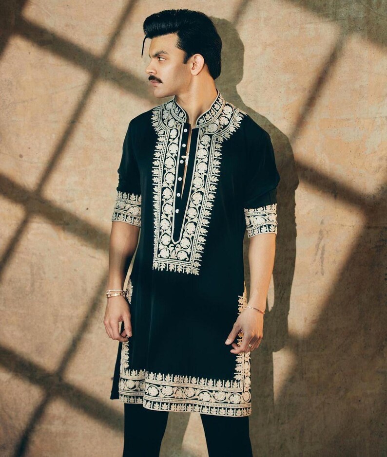 May include: Black tunic with intricate white embroidery. The tunic features a mandarin collar and button-down front. The embroidery details the collar, front, cuffs, and hem. The sleeves are three-quarter length. The person is wearing black pants.