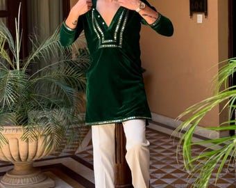 May include: A dark green velvet tunic with a V-neckline, embellished with mirrored accents, paired with cream-colored trousers. The tunic has long sleeves and a straight hem. The outfit is completed with black shoes.