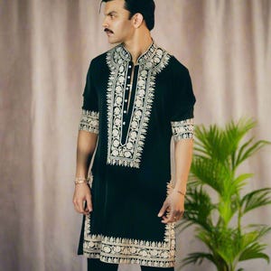May include: Black tunic with intricate gold embroidery. The tunic features a mandarin collar and decorative patterns along the front, sleeves, and hem. The garment is paired with black pants. A plant is visible in the background.
