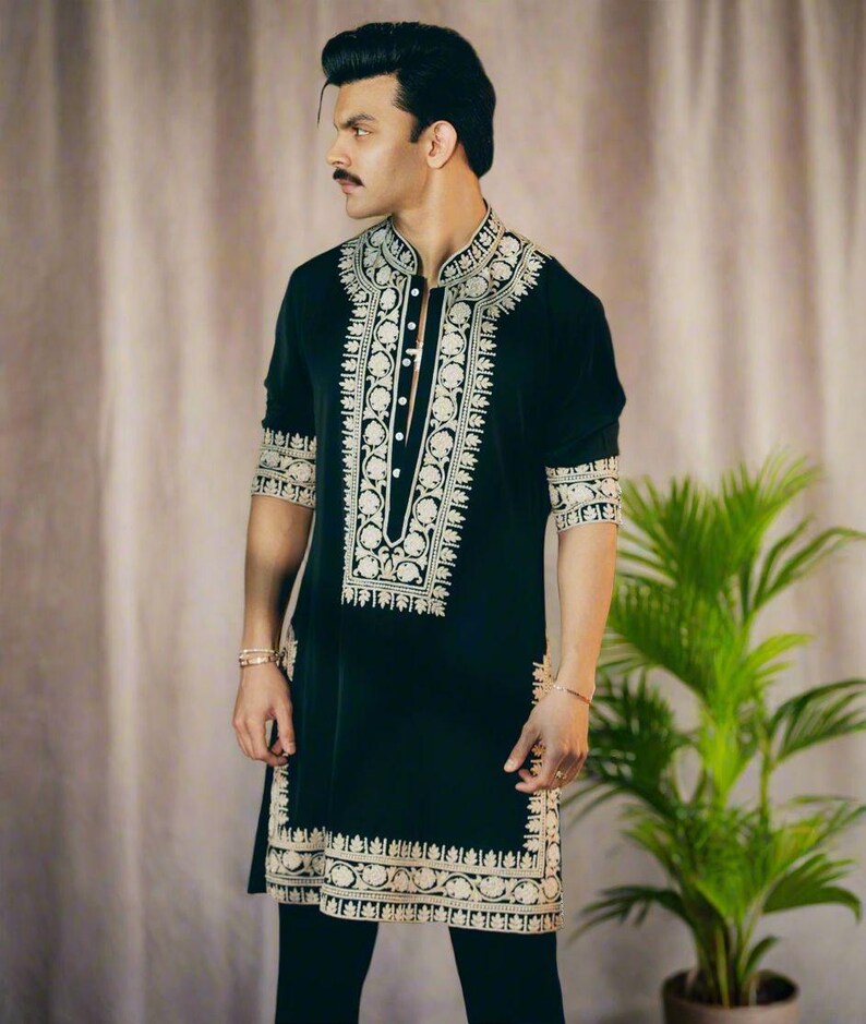 May include: Black tunic with intricate gold embroidery. The tunic features a mandarin collar and decorative patterns along the front, sleeves, and hem. The garment is paired with black pants. A plant is visible in the background.