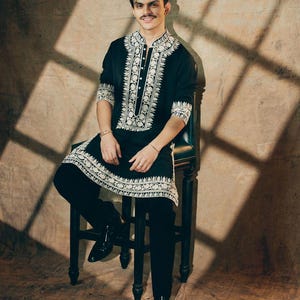May include: A black tunic and pants set with intricate white embroidery. The tunic features a mandarin collar and button-down front. The outfit is paired with black boots. The person is seated on a dark wooden chair.