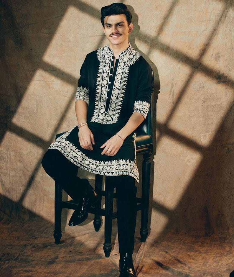 May include: A black tunic and pants set with intricate white embroidery. The tunic features a mandarin collar and button-down front. The outfit is paired with black boots. The person is seated on a dark wooden chair.