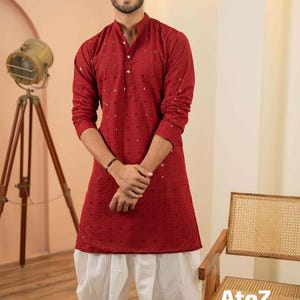 Puede incluir: Un kurta rojo con cuello mandarín y pantalones salwar blancos. El kurta presenta un patrón sutil y detalles de botones. El atuendo se combina con zapatos negros. El texto "AtoZ" es visible en una silla de madera.